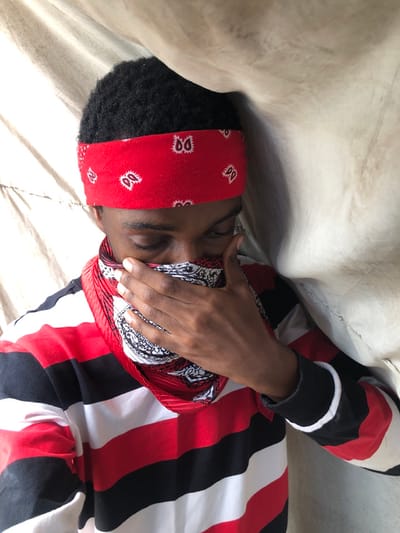 The scene is depicted in the city of Ngleshi Amanfro, Ghana. A single individual with dark skin and short, dark, tightly curled hair is positioned facing forward, with their head slightly tilted down and to the left. A red bandana with white paisley patterns is tied around their forehead. A second, multi-patterned bandana, primarily red and white with black details, covers the individual's lower face, extending from below the nose to the neck. Their right hand, with fingers slightly spread, is placed over this lower bandana, further obscuring the mouth and chin area. The individual's eyes are downcast. They are wearing a long-sleeved shirt with horizontal stripes in red, white, and black; the left cuff shows a black ribbed band with a thin white stripe. The background is composed of a light-colored, possibly beige or off-white, draped fabric.