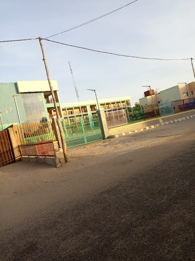 The image shows the front entrance of a building located in Maiduguri, Nigeria. The building is light green and beige with large windows. The entrance is gated with a metal fence. There is a paved road in front of the building, which leads to a small area with a few trees. The sky is clear and blue, and there are electric wires overhead. There are no people in the image. The image is taken from a slightly elevated position. The coordinates provided (latitude: 11.815, longitude: 13.155) are located in the city of Maiduguri, Nigeria.