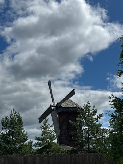 An outdoor, low-angle shot captures a traditional wooden windmill on a bright, cloudy day. The windmill, constructed from dark, horizontal logs, has a conical roof and two large, wooden blades, one of which is broken. It is partially obscured by lush green pine trees in the foreground and to the sides, as well as some lighter green leafy trees to the right. A rustic, dark wooden fence runs along the bottom of the frame, separating the viewer from the windmill and trees. The sky above is a vibrant blue, filled with dramatic, white, and gray cumulus clouds. The scene appears to be located in the city of Privolzhsky, Russia, likely in an area with historical or cultural significance, given the presence of the windmill. No people are visible in the image.
