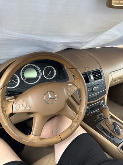 The image shows the interior of a car, likely a Mercedes-Benz, with a person's legs visible. The steering wheel is a light brown color and appears worn. The dashboard features a speedometer and tachometer. A car key is placed on the center console near the gear shifter. The interior of the car is covered with a white sheet, suggesting it might be in storage or undergoing maintenance. The scene is dimly lit, with a white sheet obscuring parts of the car's interior. The car is located in Sliven, Bulgaria.