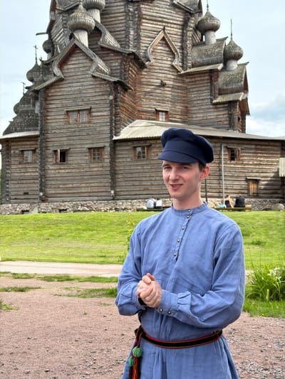 A young man wearing a blue tunic and a dark cap stands in front of a wooden church in Ust-Izhora, Russia. The man is smiling and looking at the camera with his hands clasped in front of him. The church, with its intricate wooden architecture and multiple onion domes, dominates the background. Green grass and a dirt path surround the man and the church. In the distance, other people can be seen sitting on benches. The sky is cloudy.