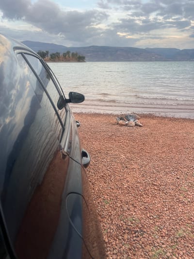 A car is parked on a reddish-brown, gravelly beach next to a body of water. The water is calm, with small waves lapping against the shore. In the distance, mountains can be seen, and the sky is cloudy. The scene is near Bin El Ouidane, Morocco.