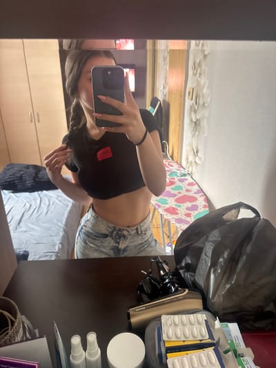 The image depicts a mirror selfie taken indoors, reportedly in Targovishte, Bulgaria. A female subject, partially visible from the chest down, is centered in the reflection. She has dark hair styled in a braid or pigtail on the left side. She is wearing a black cropped t-shirt with a red rectangular patch on the chest displaying the mirrored text "OGUH" (indicating "HUGO" on the actual garment). She also wears light-wash denim shorts. Her left arm is extended, holding a dark-colored smartphone, which obscures most of her face. Her nails are painted pink. A black elastic band is on her right wrist, and her right hand is touching her hair on the left side of her head.
The reflection of the room behind her reveals a light-colored wooden wardrobe with two doors on the left. Adjacent to it, a bed with a light blue sheet and a dark pillow/blanket is visible. To the right, an ironing board with a colorful cover featuring pink, purple, and blue-green hearts is present. The wall behind the ironing board has white, textured wallpaper or wall covering.
The foreground of the image shows a dark brown wooden surface, likely a desk or table. On the left, a woven basket with contents resembling string or yarn is visible. In the center, two white spray bottles and a white round container (possibly cosmetic items) are arranged. To the right, multiple white pharmaceutical blister packs and other medicine boxes are stacked, alongside a black plastic trash bag that is partially filled. Behind the bag, a pair of black shoes with a beige or white sole are discernible.