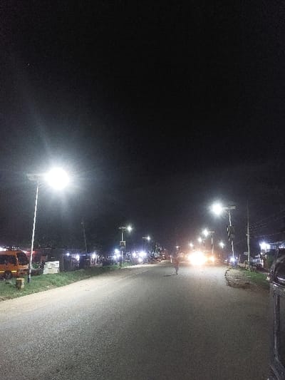 The image displays a nighttime street scene in Msata, Tanzania. A paved two-lane road, slightly curving right, extends into the distance under a dark sky. The road is illuminated by a series of solar-powered streetlights positioned along both sides.

On the left side of the road, a dirt shoulder is visible, beyond which stand illuminated structures, possibly commercial buildings or residences. An orange/yellow minibus is parked on this shoulder in the foreground.

On the right side, a similar dirt shoulder is present, with structures and parked objects, possibly motorcycles or bicycles, visible behind the streetlights. The immediate right foreground shows a partial view of a dark vehicle, indicating the vantage point.

In the center of the road, a lone individual, appearing to be an adult, walks away from the viewer, moving towards the distant bright headlights of multiple oncoming vehicles. These headlights create significant lens flare, obscuring details of the vehicles themselves. No specific interactions are discernible between the pedestrian and the approaching traffic, nor between any other individuals.