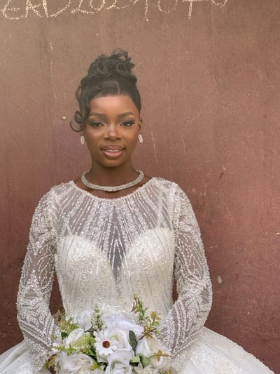 A young adult Black woman stands facing the camera, positioned centrally against a textured, reddish-brown wall. Her dark hair is styled in an elaborate updo with curls framing her face. She wears full makeup, including defined eyebrows and a subtle smile. She is adorned with a double-strand choker necklace and rectangular drop earrings, both featuring small, clear stones. She is wearing a long-sleeved white wedding gown, intricately embellished with clear beads and sequins that form patterns across the bodice and sleeves. The gown features an illusion neckline over a sweetheart cut. In her arms, she holds a bridal bouquet composed of white roses, white daisies, and various green foliage. Faint, faded white markings, possibly chalk or paint, are visible at the top edge of the wall behind her, appearing to spell "EAD 2020 TO". The scene is located in Garaku, Nigeria.