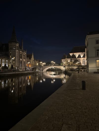 The image depicts a nighttime scene along the Leie River in Ghent, Belgium. On the left bank, the historic Graslei and Korenlei quays are illuminated by streetlights, showcasing the ornate architecture of guildhalls and other historic buildings. These buildings have numerous windows, many of which are lit from within, casting reflections on the dark water of the river. A prominent bridge, likely the Grasbrug, spans the river in the mid-ground. On the right bank, a cobblestone promenade is visible, bordered by a dark river. Further along the right bank, a large, gothic-style building, possibly a church, is brightly lit, highlighting its architectural features. A few people are visible walking on the promenade. The sky is dark, indicating it is nighttime.