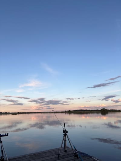 The image shows a body of water, likely a lake or wide river, reflecting the sky with clouds. The sky is a gradient of blue to orange hues, suggesting either sunrise or sunset. A shoreline with trees and vegetation is visible in the distance. In the foreground, there is a wooden pier or dock with fishing equipment present, including a rod stand, fishing rods, and a landing net. The location is likely Khryashchevka, Russia. The overall scene portrays a tranquil and recreational setting, possibly involving fishing or relaxation by the water.