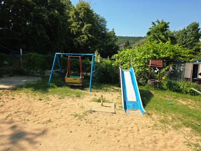The image depicts a small, somewhat overgrown playground located in the city of German, Bulgaria. The playground area is covered in sand, with patches of grass. Prominently featured are a blue and red metal swing set with a multi-colored seat, and a blue metal slide with white and blue markings. In the background, dense green foliage and trees dominate the scene. To the right, a small, rustic structure with a red corrugated metal roof, possibly a shed or playhouse, is visible. The sky is clear and blue, indicating a sunny day. No people are present in the image. The overall impression is that of a simple, community-based outdoor recreational space.