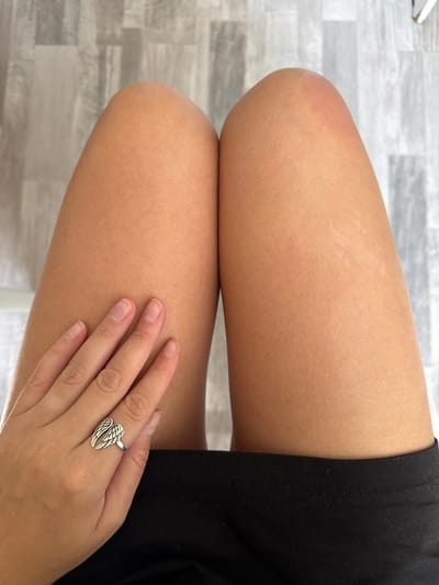 This is a close-up, eye-level shot of a person's thighs and hand. The person is wearing black clothing at the bottom of the frame, and their skin appears tanned. A hand with unpainted nails is resting on the left thigh, adorned with a silver ring shaped like angel wings. The background is a textured, light-colored surface that resembles wood flooring. The image was captured in Cheboksary, Russia.