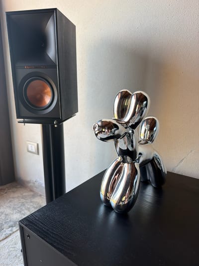This is a medium shot, eye-level, indoor photograph taken in Asciano, Italy, featuring a black wooden table with a chrome balloon dog sculpture and a black speaker on a stand. The chrome balloon dog sculpture is in the foreground, positioned on the right side of the table, with its reflective surface mirroring the surrounding environment. To the left, a black speaker is mounted on a black stand. The speaker has an orange woofer and a horn-shaped tweeter. A light switch is visible on the wall behind the speaker. The background consists of a textured, light-colored wall and a portion of a grey floor. The lighting is soft, casting subtle shadows.