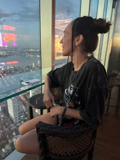 A person sits in a chair at a table by a large window, overlooking a city at dusk. The person, with dark hair in a bun and braids, wears a dark t-shirt with a graphic print and a necklace. They are looking out the window at the city lights and the sky. On the glass table in front of them is a phone. The city appears to be Hanoi, Vietnam, based on the distinctive architectural style visible in the background and the presence of the Lotte Center Hanoi, a prominent skyscraper in the city. The perspective from a high vantage point suggests the location is likely a bar or restaurant on an upper floor of a building.