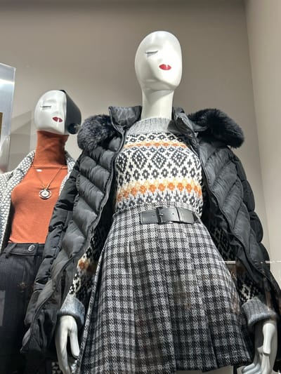 This image captures a retail display featuring two mannequins showcasing winter fashion ensembles. The scene is static, likely within a store or display window in Koridallos, Greece.
The prominent mannequin, positioned slightly higher, is depicted standing upright. It wears a black puffer jacket with a faux fur trim around the hood or collar, unzipped to reveal a layered outfit. Beneath the jacket is a grey sweater featuring a distinctive Fair Isle or Nordic-inspired pattern in white, black, and burnt orange or brown hues. A grey and black houndstooth or small check patterned skirt is worn on the lower body, cinched at the waist with a black belt and rectangular buckle. The mannequin itself is white with simplified facial features and striking red "eyes."
Partially visible below the first is a second mannequin, also standing. This mannequin wears a terracotta or burnt orange turtleneck sweater, accessorized with a dark cord necklace featuring a circular, silver-toned pendant. A black puffer jacket is layered over the turtleneck, and dark-colored pants or jeans are visible below. Another black and white geometric patterned garment peeks out from beneath the puffer jacket. This mannequin also has white skin and similar red "eyes."
The background is a plain light grey wall, suggesting a clean display environment. The lighting is uniform, highlighting the textures and patterns of the clothing. There is no visible text in the image.