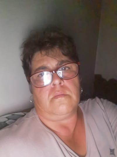 The image is a selfie of a woman in Lomets, Bulgaria. The woman has dark, curly hair and is wearing glasses with red frames. She is also wearing a light-colored v-neck shirt. The background is dark and out of focus. The woman is looking directly at the camera with a neutral expression. The image appears to have been taken indoors, possibly in a home.