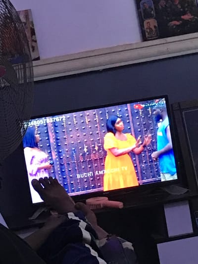 A television displays a program featuring two individuals on a patterned carpet. One person wears a yellow dress and the other is in blue. The screen displays "BUCHI AMAECHI TV" and a phone number. A foot rests near the screen. In the foreground, a fan is partially visible. The overall image appears casually taken indoors.