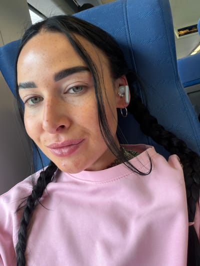 The media file shows a close-up, vertically oriented shot of a woman with dark hair in a braid, wearing a pink sweatshirt. She has light-colored eyes, prominent eyebrows, and visible freckles on her face. She is wearing a white earbud in her right ear, which has the branding "HUAWEI" visible. She also has a small, sparkly earring in her ear. Her lips appear to be glossy.
The woman is seated on a blue fabric surface, which appears to be a seat with a patterned texture. In the background, a yellow sign with black text is visible, reading "135km/4". This suggests a transportation setting, possibly a train or bus. The lighting indicates it is daytime. The overall mood is casual and relaxed.
The location context provided is Ушаки, Russia.