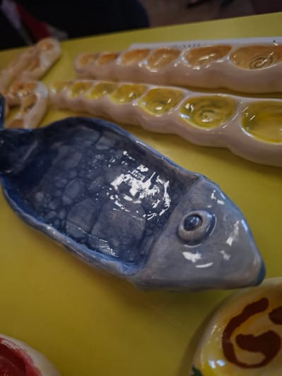 A close-up, high-angle, eye-level shot shows a collection of ceramic dishes arranged on a bright yellow surface. In the foreground, a dark blue ceramic dish shaped like a fish is prominently displayed. The fish dish has a textured, slightly irregular surface with visible brush strokes and a single eye. Behind the fish, two lighter-colored ceramic dishes with multiple indentations are visible. These dishes appear to be shaped like long, narrow loaves, possibly bread, with yellow and orange glaze within the indentations, some with swirling patterns. In the lower right corner, a portion of another ceramic object with brown and yellow markings is partially visible. The background is dark and out of focus, suggesting an indoor setting. The overall impression is of a display of handmade ceramic crafts. The image does not provide any specific information or visual cues that would allow for the determination of its location within the city of Vidin, Bulgaria.