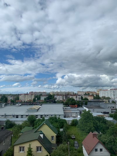 The image captures a cityscape of Kaliningrad, Russia, under a cloudy sky. In the foreground, several residential houses with prominent pitched roofs are visible, some painted yellow and others gray. These houses are surrounded by lush green trees and foliage. A large, light-gray industrial building with a corrugated metal roof dominates the mid-ground, featuring multiple windows. In the distance, a cluster of tall, multi-story apartment buildings, typical of Soviet-era architecture, stretches across the horizon. An orange bus is partially visible among the industrial structures. The sky is filled with dramatic, heavy clouds interspersed with patches of bright blue sky. The overall scene suggests a typical urban environment with a mix of residential and industrial areas, set against a backdrop of a dynamic sky.