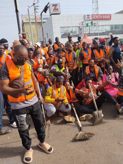 The image depicts a large group of approximately 30-40 individuals, primarily young adults, gathered on a paved urban street in Fagge C, Nigeria. The background features a multi-story white building prominently displaying the "Zenith Bank" logo and signage, including a large 'Z' and 'ZENITH' text, along with Nigerian flags. Another building with "aoju" visible text is present further down the street, beneath overhead power lines and a traffic signal structure.
Most individuals in the group are wearing bright orange reflective safety vests, some with "KANO" text. Many are holding cleaning tools, specifically traditional brooms made of bundled sticks. One man seated in the lower right holds a shovel. Several people are wearing white gloves. The group is largely clustered, posing for the camera with various expressions, including smiles, peace signs, and thumbs-up gestures.
In the foreground left, a man wearing a black t-shirt, orange vest, dark sunglasses, and light-colored slip-on sandals stands, holding a white-handled broom and looking down at a smartphone in his right hand. A brown sling bag is worn across his chest. Two women crouching in the mid-ground also appear to be looking at their phones. The ground is a light gray asphalt surface, and the sky is bright and clear, indicating daylight.