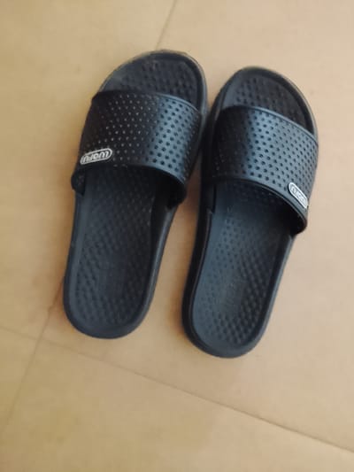 A pair of black "LUOFU" brand slide sandals are placed side-by-side on a light brown, tiled surface. The sandals are made of a matte material with a perforated strap across the top of each footbed. The footbeds themselves have a textured, bumpy surface, and the soles appear to be thick and black. The lighting is moderate, casting faint shadows on the floor. The overall scene is static and appears to be an indoor setting. The image does not provide any information about the city of Sunkani, Nigeria.