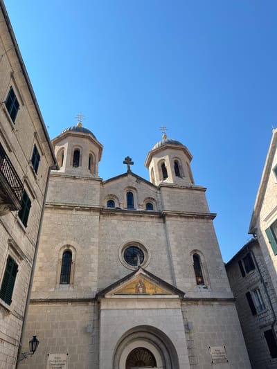 The image shows the Church of St. Nicholas in Kotor, Montenegro, viewed from a low angle looking up. The church's facade is constructed of light-colored stone and features two bell towers topped with domes and crosses. The sky above is a clear, bright blue. To the left and right of the church are tall, narrow buildings, also made of stone, with windows and shutters. The building on the left has a wrought-iron balcony. A traditional lamppost is attached to the wall of the church on the lower left. The image is well-lit, suggesting it was taken on a sunny day.