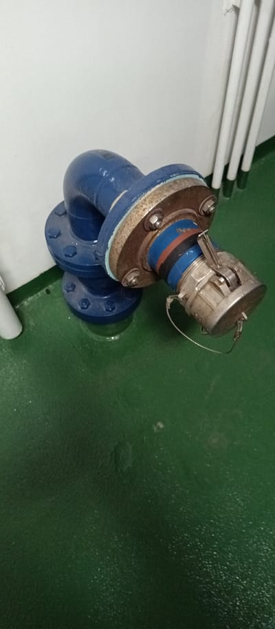 A blue pipe elbow with flanges is attached to a metal coupling. The pipe is on a green floor, with a white wall and white pipes in the background. This appears to be located in Al Hamriyah, United Arab Emirates.