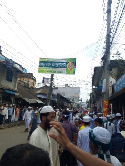 The image depicts a bustling street scene in Bhola, Bangladesh. The location is identified by its coordinates (latitude: 22.686, longitude: 90.644). The street is crowded with people, predominantly men, many of whom are wearing traditional white garments. There is a large banner hanging above the street, written in Bengali. Several shops are lined along the sides of the street, with a variety of goods displayed. An electric pole with numerous cables extends overhead, adding to the chaotic yet lively atmosphere of the scene. The focus is on the crowd of people, their movement, and the overall ambiance of the street.