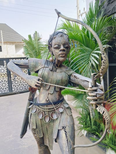This is a full-body, eye-level, outdoor shot of a life-sized metal sculpture of a female archer in what appears to be Dutsen Alhaji, Nigeria. The sculpture is positioned centrally in the frame, facing forward with her body angled slightly to her left. She is depicted in a dynamic pose, drawing back a bow with a string taut, as if preparing to shoot an arrow. Her left arm holds the bow steady, while her right hand is drawing the string towards her face.
The sculpture is made of what appears to be aged metal, with a greenish-bronze patina suggesting weathering. She is adorned in detailed armor or decorative metal plates that cover her torso and legs, resembling segmented scales. Circular metal adornments hang from a belt around her waist, and she wears a quiver filled with arrows on her back. Her facial features are distinctly rendered, with closed eyes and a focused expression. She wears earrings and appears to have eye makeup or adornments.
Behind the sculpture, the environment consists of lush green tropical foliage, primarily palm fronds and ferns, suggesting a warm climate. To the left of the sculpture, a section of a decorative metal gate with an intricate geometric pattern is visible, hinting at an entrance or boundary. Further in the background, a building under construction, with visible brickwork and a dark-tiled roof, is partially obscured by the vegetation. Overhead, power lines stretch across the sky, indicating an urban or semi-urban setting. The ground beneath the sculpture is paved with what appears to be concrete or stone slabs. The lighting is natural, indicating it is daytime, likely with overcast conditions given the diffused light.