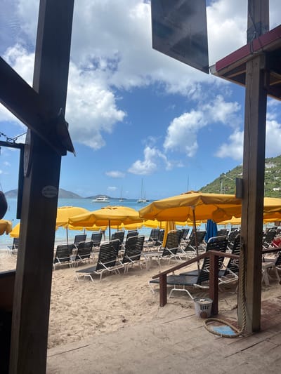 The image depicts a beachfront scene in Road Town, British Virgin Islands, observed from a shaded elevated wooden structure. The foreground features dark brown wooden support beams and a partial reddish roof overhang. On the wooden deck, a white Miller Lite branded bucket filled with sand serves as an ashtray, and a coil of rope with a blue-painted base rests beside a wooden railing.

Beyond the wooden structure, a light sandy beach extends towards the water. The beach is populated with numerous rows of unoccupied black and white striped lounge chairs, each paired with a bright yellow beach umbrella providing shade. The umbrellas form a continuous line along the beach.

The calm, clear turquoise ocean occupies the midground, with several boats anchored or sailing. These include multiple white sailboats and at least one larger white catamaran or yacht. In the distant background, lush green hills rise from the water, dotted with scattered white and light-colored buildings. The sky above is a clear blue with scattered white cumulus clouds. No explicit human activity is clearly discernible, though a red garment is faintly visible near the lounge chairs on the far right.