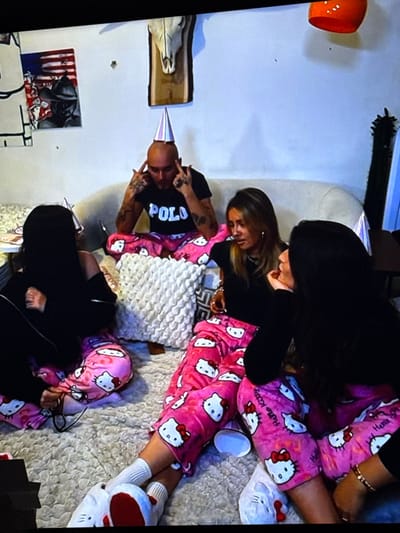 Four individuals, one male and three females, are depicted indoors in what appears to be a casual social gathering. The specific location within the city of Vellmar, Germany, is not visually identifiable in the image.
The scene features light-colored walls, a light-colored sofa, and a fluffy white rug or blanket covering the floor area. Wall decor includes a framed artwork depicting an American flag motif with a dark figure, a bone skull mounted on a wooden plaque, and a partially visible orange, bowl-shaped object. A tall, dark, spiky decorative object is visible on the far right.
All four individuals are dressed in pink pajamas patterned with white "Hello Kitty" characters. Three individuals are wearing silver conical party hats.
The male, positioned centrally and seated on the sofa, is bald and wears a black t-shirt with "POLO" written in white. He has visible tattoos on both forearms. He is wearing a party hat and has both index fingers pointed towards his temples in a distinct gesture.
To the male's left, a female sits on the floor, partially obscured, wearing a black top and a party hat. Dark-rimmed glasses are resting on her lap.
To the male's right, a female sits on the floor, wearing a black top. She is looking towards the female on the far right. She is not wearing a party hat. Her lower legs are visible, showing white socks and white "Hello Kitty" slippers.
On the far right, a female sits on the floor, wearing a black top and a party hat. She is looking towards the female in the middle, with one hand near her chin in a conversational posture. A gold bracelet is visible on her left wrist.
A white, textured decorative pillow rests on the floor between the seated individuals. A white, circular object is on the rug. A dark table or surface is partially visible on the far left, holding some indistinct items.