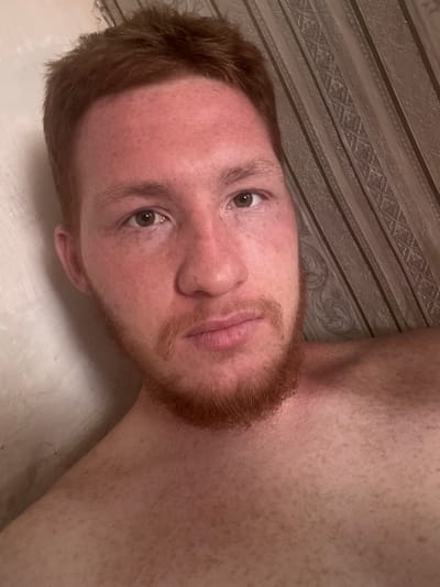A close-up, high-angle, selfie-style photograph shows a fair-skinned individual with reddish-brown hair and beard, facing the camera. The person appears to be indoors, with a lightly textured, off-white wall on the left and a striped, patterned wallpaper in muted brown tones on the right. The subject's bare chest is visible, showing freckles. The overall lighting is dim, with some reddish tones suggesting artificial light. There are no clear indications of the specific location in Makiivka, Ukraine, beyond the subject being in an indoor environment.