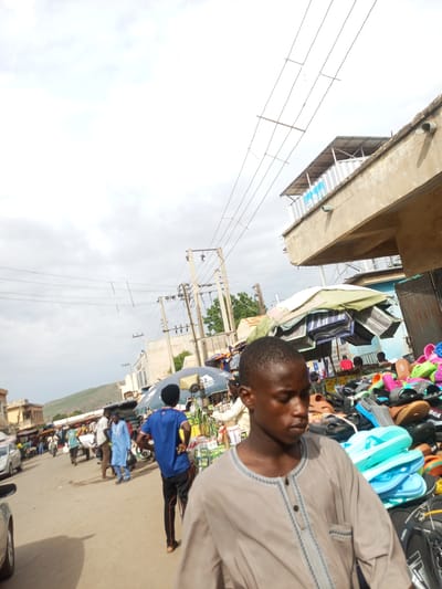 This image depicts a street market scene in Arawa, Nigeria. In the foreground, a young man wearing a light brown, traditional tunic with gray trim is looking down, his face partially obscured. To his right, a large display of various colorful sandals and flip-flops is visible, with stacks of blue and green footwear prominent. In the middle ground, a man in a bright blue tunic and black pants is walking away from the camera, his hands on his hips. Behind him, another man in a traditional white robe and brown pants is walking. A motor scooter is parked nearby. The street is unpaved and dusty. In the background, there are stalls with umbrellas and awnings, and more people are visible. Buildings with concrete structures and metal roofs line the street, and electrical poles with numerous wires are present. A hillside is visible in the far background. The sky is overcast with visible power lines crisscrossing overhead.
