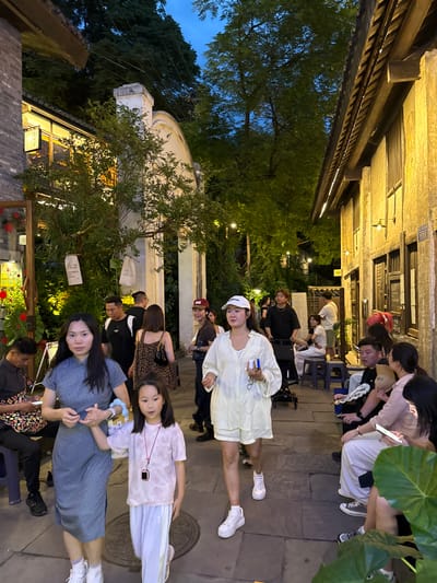 A nighttime scene in Nan'an District, China, shows a narrow cobblestone street illuminated by warm lighting from buildings and strung lights. A diverse group of people are gathered, primarily seated on small stools, suggesting an outdoor dining or social setting. Individuals appear to be engaged in conversation or waiting, some glancing at the camera. Plant life is visible throughout, adding to the ambiance, and the architecture is a mix of traditional and modern styles.