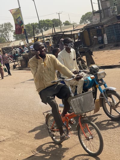 The image captures a daytime street scene in Chikaji, Nigeria, under bright sunlight. In the immediate foreground, a young adult male, with dark skin and a light beard, wears a light beige hooded sweatshirt and dark grey trousers. He is positioned on an orange folding bicycle, holding a dark smartphone to his right ear and smiling, looking towards the left. A silver-colored slatted basket is affixed to the front of his bicycle. Adjacent and slightly behind him, an adult male, dressed in light-colored traditional attire, glasses, and a camouflage-patterned face mask, sits on a light blue motorcycle, gripping the handlebars and facing forward. A child in a light-colored top is partially visible between the two men.
The background shows a populated street with multiple individuals, some walking, others standing or gathered near shaded structures. Several parked motorcycles are visible. Buildings with corrugated metal roofs and walls line the street, indicative of commercial establishments or market stalls. A partially visible sign on one building reads "GDI SUPERSTORE ELECTRICAL AND MECHANICAL SPARE PARTS". A tall utility pole featuring a colorful red and yellow banner stands prominently to the left of the center. The ground consists of a light brown/grey asphalt or dirt road, exhibiting distinct shadows cast by the subjects and vehicles. The sky is hazy and light blue.