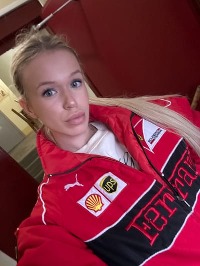 A young Caucasian woman with blonde hair styled in a braid and light blue eyes is looking directly at the camera, taking a selfie. She is wearing a red jacket with white and black accents, featuring embroidered logos of "Scuderia Ferrari," "UPS," and "Shell." The jacket also has a Puma logo. She is also wearing a cream-colored sweater underneath. The background is mostly out of focus, but appears to be indoors with a red wall and some wooden elements, possibly a hallway or room. The location is described as the city of Shushary, Russia.