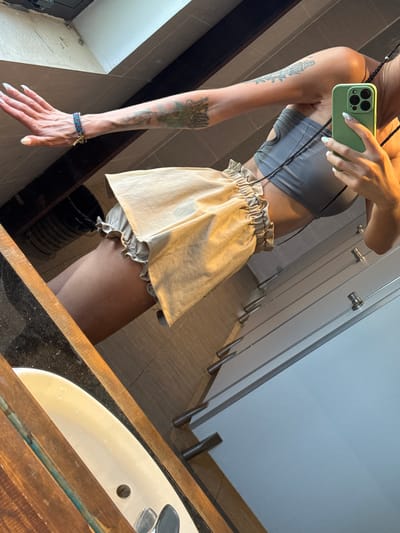 A person is seen taking a mirror selfie in what appears to be a public restroom, located in Hà Giang 1 Ward, Vietnam. The image captures a partial reflection of the individual, focusing on their torso, left arm, and upper legs.
The person is wearing a grey ribbed crop top and a light beige or tan high-waisted ruffled skirt, with ruffled undergarments or shorts visible beneath the skirt. They are holding a light green smartphone with a black camera module in their right hand, reflected as their left, positioned to capture the image. The phone has a black strap, possibly worn around the neck or shoulder. The person's left arm is prominently displayed, adorned with multiple tattoos: a detailed, tower-like structure with flowing vines or leaves on the upper arm, and a darker, less decipherable design on the lower forearm. A multicolored beaded bracelet with a small charm is visible on their left wrist. Their nails are long and manicured with a light, possibly iridescent, polish.
The background of the reflection reveals a row of several light-colored, closed stalls or cabinets, each with a silver handle or pull. In the immediate foreground, part of a white sink with a drain and a metallic faucet is visible, set into a dark countertop with a rustic-looking wooden frame around the mirror and sink area. A black wicker-like basket or trash can is faintly reflected on the floor beneath the skirt. The lighting suggests an indoor setting, likely daytime. No discernible text is visible on any objects or surfaces in the image. The activity is clearly a mirror selfie.