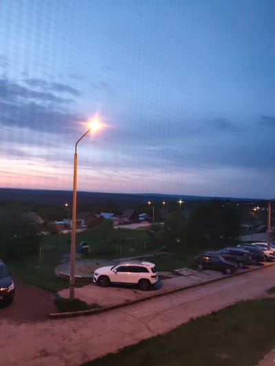 The image depicts a dusk scene in Gremyachinsk, Russia. In the foreground, a street lamp illuminates a parking area along a sidewalk. Several cars are parked, including a white SUV prominently positioned near a playground with a sandpit and slide. Beyond the parking area, the landscape descends to reveal rooftops of houses in the lower ground of the city. In the background, there's a dark, dense forest, possibly marking the edge of the inhabited area. The sky is a gradient of colors, transitioning from orange and pink near the horizon to blue and gray towards the upper part of the image, suggesting either sunrise or sunset. Other street lamps are sparsely distributed in the distance, indicating other residential areas. The area appears residential and quiet.
