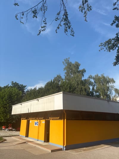 A low-angle shot captures a bright yellow, modern public restroom building under a clear blue sky with a few wispy clouds. The building has a white overhang, and the yellow facade is divided into at least two distinct sections. One section features a blue and white "WC" sign, and next to it, a yellow sign with white text reads "Herren" (Men's). Below the "Herren" sign, another yellow sign with white text reads "Damen" (Women's). A light blue trim runs along the base of the yellow facade.
Lush green trees form a dense backdrop to the building on the left side of the image, with their branches and leaves reaching into the sky. On the right side, a sidewalk or paved area is visible, leading away from the building. In the background, some red chairs are arranged on a stone or gravel surface, suggesting an outdoor seating area. The sunlight indicates it is daytime, likely a sunny day given the clear sky and the bright appearance of the building and foliage. The scene appears to be a public space, possibly a park or a rest stop, given the restroom facility and seating. There is no visible activity taking place. The overall mood is peaceful and bright.