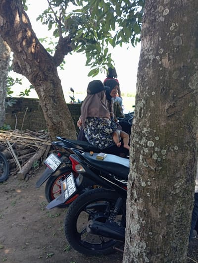 A woman wearing a headscarf is holding a baby. A motorcycle with license plates S 3959 BCZ and S 2523 BOE is parked next to them. A tree is in the background. The photo was taken in the city of Waung, Indonesia.