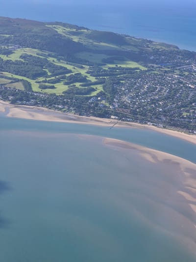 An aerial view captures a coastal landscape with a town nestled between a body of water and a green, hilly terrain. The foreground is dominated by a large expanse of calm, blue-green water, with visible sandbanks or shallow areas. Beyond the water lies a sandy beach, bordering a developed area. This area consists of a dense collection of houses with light-colored roofs, typical of a suburban or coastal town. Behind the residential zone, the landscape transitions to lush green fields, suggesting parkland or agricultural use, interspersed with scattered trees. Further inland, the terrain rises into rolling hills covered in darker vegetation, possibly heathland or dense woodland. The sky above is a clear, pale blue, indicating fair weather. The scene appears to be a peaceful, residential coastal area, likely on the outskirts of a city or a large town. Based on the landscape and typical coastal development in Ireland, this could be an area like Howth, Dublin, Ireland, known for its hilly coastline, residential areas, and proximity to the sea.
