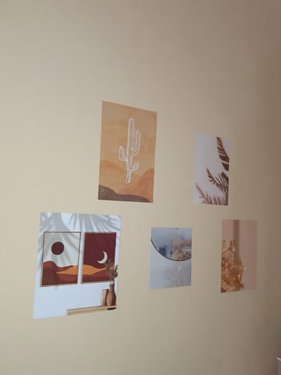 The image depicts a wall decorated with several framed art prints. The wall is painted a light, neutral color, possibly beige or cream.

There are five art prints arranged on the wall.
On the upper left, there is a print with a stylized white cactus on a background of earthy tones, suggesting a desert landscape.
To its right, a smaller vertical print features the silhouette of a palm frond or similar foliage.
Below the cactus print, a larger horizontal print displays two framed scenes. The left frame shows a dark red sun over abstract desert hills, while the right frame shows a crescent moon over similar hills, with a small vase of dried flowers placed in front of it on a wooden shelf.
To the right of the two desert scenes, and slightly below the palm frond print, there is a small circular mirror with some dried flowers visible within its reflection.
Finally, to the right of the mirror and the palm frond print, there is another small vertical print with an image of dried flowers against a soft pink background.

The lighting suggests an indoor environment, with natural light casting shadows from the framed prints and a nearby object not fully visible in the frame. The overall aesthetic is minimalist and bohemian, with a focus on natural elements and warm, earthy colors.

The location of Lebbae, Indonesia is not visually evident from the image itself. The image focuses solely on the interior wall decoration.