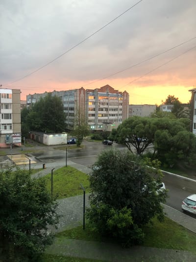 The image shows a residential area in the city of Solikamsk, Russia, during sunset. The sky is a mix of grey clouds and orange and yellow hues from the setting sun. In the foreground, there are several trees and a paved walkway. In the middle ground, there is a street with cars, a small white building with the inscription "ШИНОМОНТАЖ" (tire fitting service), and garages. In the background, there are multi-story apartment buildings. One building, located centrally, has its windows reflecting the sunset colors. Another apartment building to the left has a sign with the word "МАЯК" (Mayak). The overall scene depicts a typical evening in a Russian city.