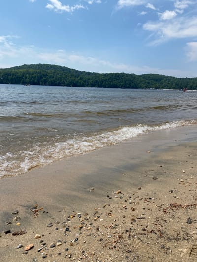 The image depicts a serene lakefront scene in Duhamel, Canada, on a clear, sunny day. The foreground is dominated by a sandy beach littered with small pebbles and seashells, with a gentle wave lapping at the shore. In the mid-ground, the calm lake stretches out, reflecting the blue sky and scattered white clouds. Along the distant shoreline, a lush green forest covers rolling hills, with a few buildings and docks visible amongst the trees. Several boats, including a kayak with two occupants and a stand-up paddleboard with two individuals, are scattered across the water, indicating recreational activity. A few distant figures can be seen on the far beach, enjoying the summer weather.