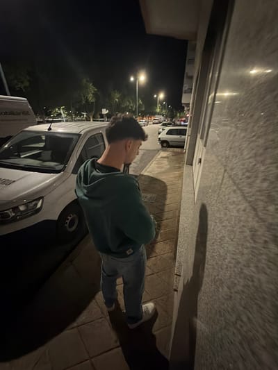 The image shows a man standing on a sidewalk at night in Bragança, Portugal. He is wearing a green hoodie, jeans, and white sneakers. He is standing next to a white van parked on the street. Streetlights illuminate the scene. There are other parked cars and buildings visible in the background. The wall of a building is on the right side of the frame, with the man's shadow cast on it.