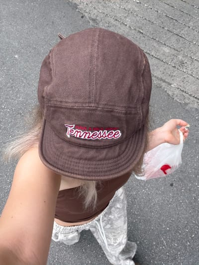 The image shows a person from the chest up, wearing a brown baseball cap and a brown tube top. The cap has the word "Tennessee" embroidered in red and white on the front. The person is holding a white plastic bag with a red object inside. They are standing on an asphalt surface, likely a street or sidewalk, with a textured pattern of light and dark gray. Their hair is blonde and appears to be somewhat messy, falling from under the cap. The overall scene is casual and appears to be taken outdoors during the daytime. The location is not specified in the image.