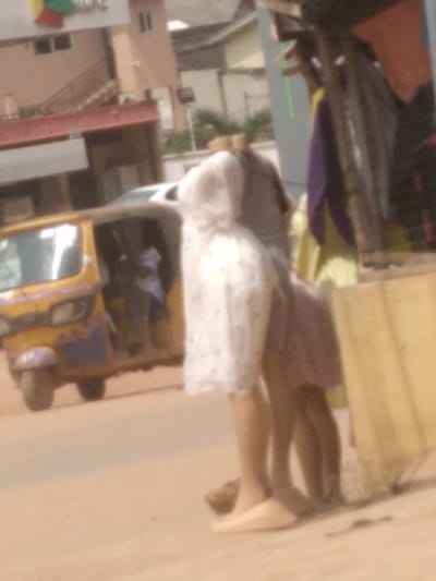 This image captures a street scene in Agege, Nigeria. In the foreground, positioned to the right, two individuals are partially visible, exhibiting a soft focus. One individual wears a light-colored, possibly white, patterned dress reaching mid-thigh, complemented by light-toned flat shoes. Adjacent and partially behind, a second individual is discernible in a light purple or pinkish dress. They are situated next to a structure constructed with wooden supports, from which various colored fabrics—including purple, yellow, and orange—are suspended, indicative of a market stall. The ground in this area is unpaved and appears as a light brown surface.

In the midground, to the left, a yellow and blue Keke Napep (auto-rickshaw) is present, with two indistinct figures visible within its passenger compartment. A white vehicle is partially visible behind the Keke Napep. The background reveals multiple buildings: one features a prominent multi-colored geometric sign (red, green, blue, yellow) on its upper facade, and another building exhibits a reddish hue with an external staircase. The environment suggests an active urban or commercial setting.