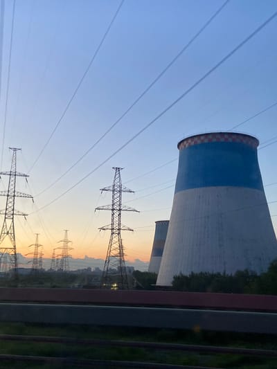 The image captures a view of a power plant at dusk in Shushary, Russia. Two large cooling towers dominate the scene, one prominently in the foreground, painted blue and white with a checkered rim. In the background, multiple high-voltage electricity pylons are silhouetted against the soft orange and blue hues of the sunset. The sky is clear, with faint contrails from aircraft streaking across it. Vegetation and a distant cityscape are visible between the pylons and the cooling towers. A dark foreground element, possibly a road or railway, runs horizontally across the bottom of the frame. There are no people visible in the image.