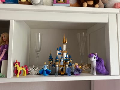 The image displays a white shelf in what appears to be a room, likely a child's bedroom given the collection of toys. The shelf is divided into sections, and the primary focus is on a display of various figurines and a Lego castle.
In the left section, a Barbie doll with blonde hair and a purple dress is visible. Next to her, on the shelf, is a small yellow and pink toy horse with a red jewel on its flank, and a silver tiara with pearls.
The central section features a Lego castle, primarily gray with blue and gold accents, resembling a fairytale castle. In front of the castle, from left to right, are small figurines of Cinderella in a blue dress, a clear blue horse-like figure, and Rapunzel in a purple dress. To the right of Rapunzel is a golden, leaf-shaped decoration.
The right section contains two clear glass flutes and a white toy horse with a purple mane and tail, adorned with a pink and purple headband.
The overall setting is a well-lit indoor environment, with the toys neatly arranged on a white shelf. The subjects are toys, including dolls, toy horses, figurines, a Lego castle, and decorative items. No specific activity is taking place; the scene is a static display. There are no visible text elements in the image. The time of day and weather are not discernible.