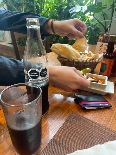 The image depicts a close-up view of a table at an outdoor restaurant, likely in Cuautitlán Izcalli, Mexico, with lush green foliage in the background. A glass of dark beverage with ice is prominently featured in the foreground, next to a glass bottle of Coca-Cola. The bottle's label displays nutritional warnings in Spanish, indicating "EXCESO CALORIAS" (Excess Calories) and "EXCESO AZUCARES" (Excess Sugars).

In the middle ground, a hand is shown reaching into a basket of tortilla chips and bread. Beside the basket is a white dish containing a dipping sauce and a slice of pickled jalapeño pepper. A small wooden utensil is being used to scoop the sauce. To the right of the dipping sauce, a small bottle of hot sauce with a red label is visible. A black wallet with colorful card slots is placed on the table near the dipping sauce.

The table is made of wood, and a textured brown placemat is partially visible in the lower right corner. The overall scene suggests a casual dining experience, possibly involving appetizers or snacks. The presence of specific food items and the Coca-Cola bottle with Mexican labeling strongly indicate a Mexican setting. The natural greenery in the background implies an open-air or well-lit interior space, perhaps a garden restaurant.