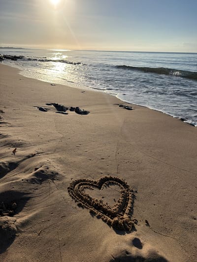 The image depicts a tranquil beach scene at sunrise in Mont-roig del Camp, Spain. A heart shape has been drawn in the wet sand near the waterline. Gentle waves are lapping at the shore, and the sun is low in the sky, casting a warm, golden light and creating reflections on the water. The sand is a light brown color, with some ripples and footprints visible. In the distance, the coastline stretches out, and the sky is a pale blue. There are no people visible in the frame.