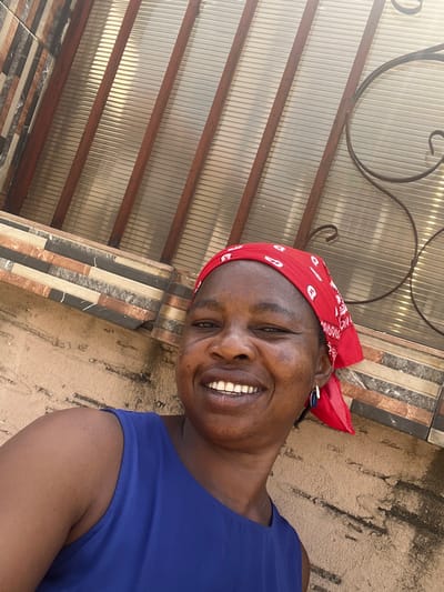 An adult female is depicted in a medium-shot, facing directly towards the camera with a broad smile, her teeth visible. She has a medium complexion and wears a red bandana with white patterns covering her head, a royal blue sleeveless top, and a dark blue teardrop-shaped earring with a white accent on her right ear. The background, from bottom to top, features a tan textured wall, followed by a horizontal band of multi-colored rectangular tiles in brown, beige, and black hues. Above the tiles, a translucent corrugated or ribbed structure, possibly a window or awning, is visible, supported by diagonal dark-colored bars. Part of a decorative dark metal scrollwork, consistent with security grating, is present on the upper right. The scene is brightly lit, indicative of an outdoor environment in Iranla, Nigeria.